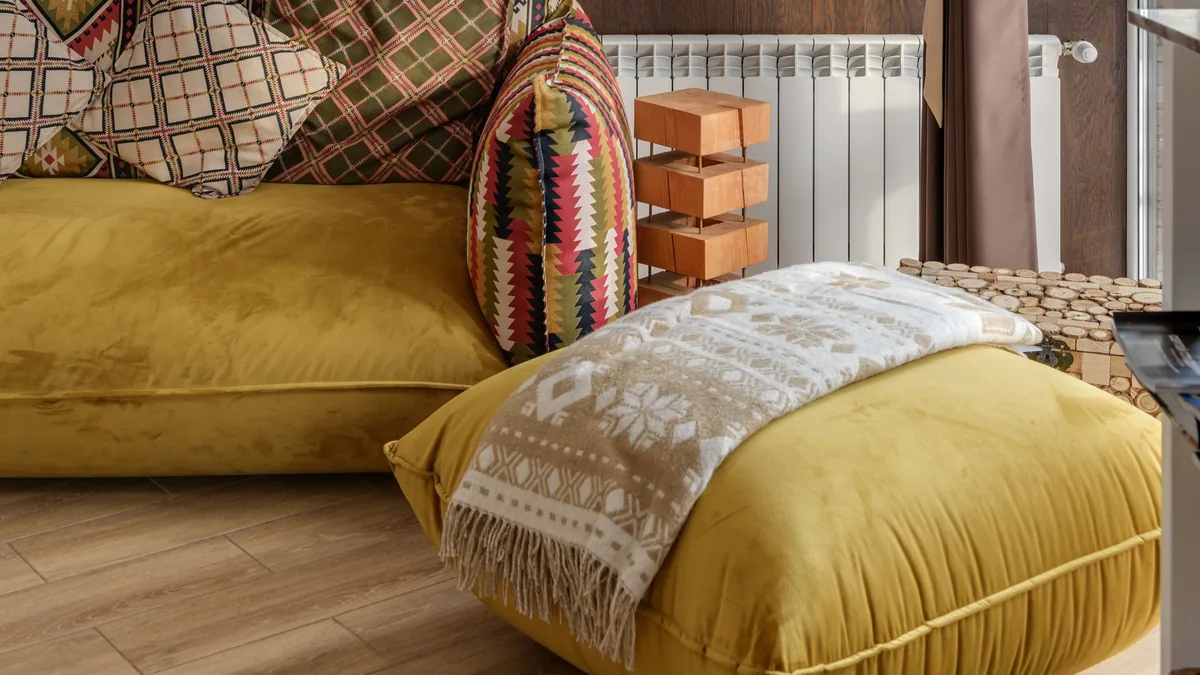 Sofá e pufe em amarelo manteiga com almofadas estampadas, manta clara e piso de madeira.