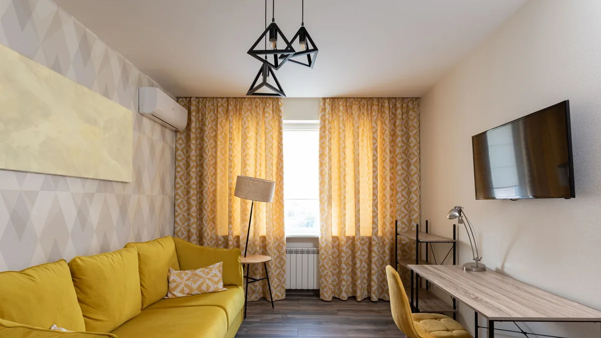 Sala pequena com sofá amarelo, cortina estampada clara, parede suave e mesa de trabalho lateral.