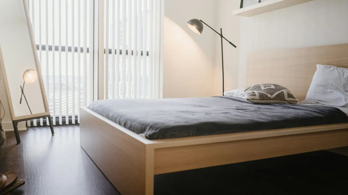 Quarto pequeno com cama de madeira clara, roupa de cama cinza, luminária de piso e janela com cortina translúcida.