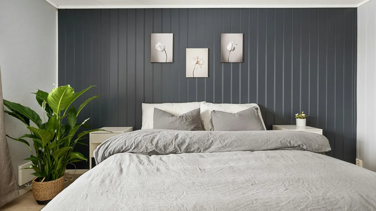 Quarto pequeno com parede cinza escura equilibrada por cama clara, planta grande e mesas laterais brancas.