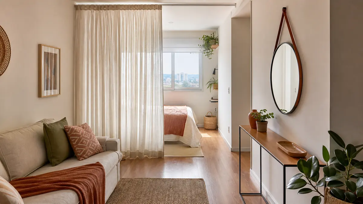 Cortina de linho claro separando a cama de uma sala compacta, com janela livre, sofá e caminho de circulação visível.