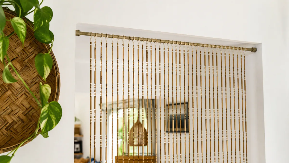 Detalhe de cortina de bambu instalada em suporte removível no alto de um vão branco com planta ao lado.