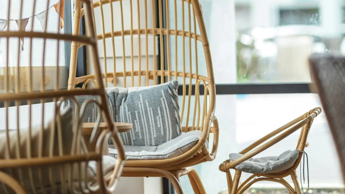 Cadeiras de rattan perto de janela com plantas suspensas, almofada clara e luz natural.