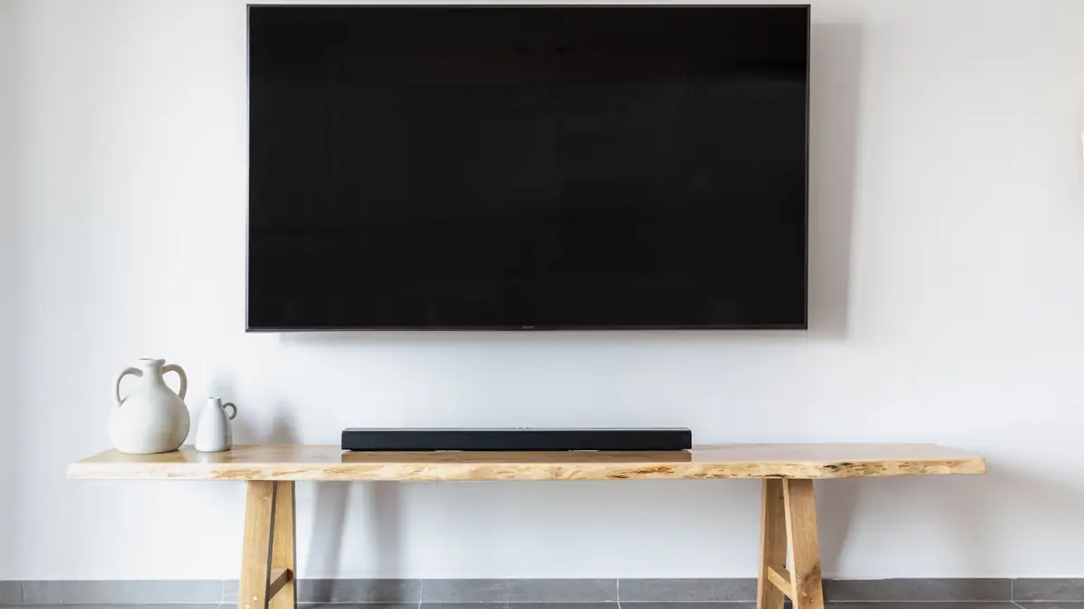 TV grande instalada em parede branca sobre mesa de madeira clara, com soundbar centralizada e poucos objetos.