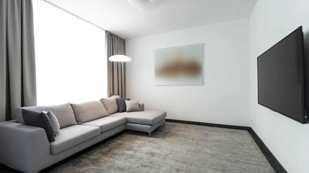 Sala clara com TV lateral na parede branca, sofá cinza, cortina longa e luminária pendente acesa.