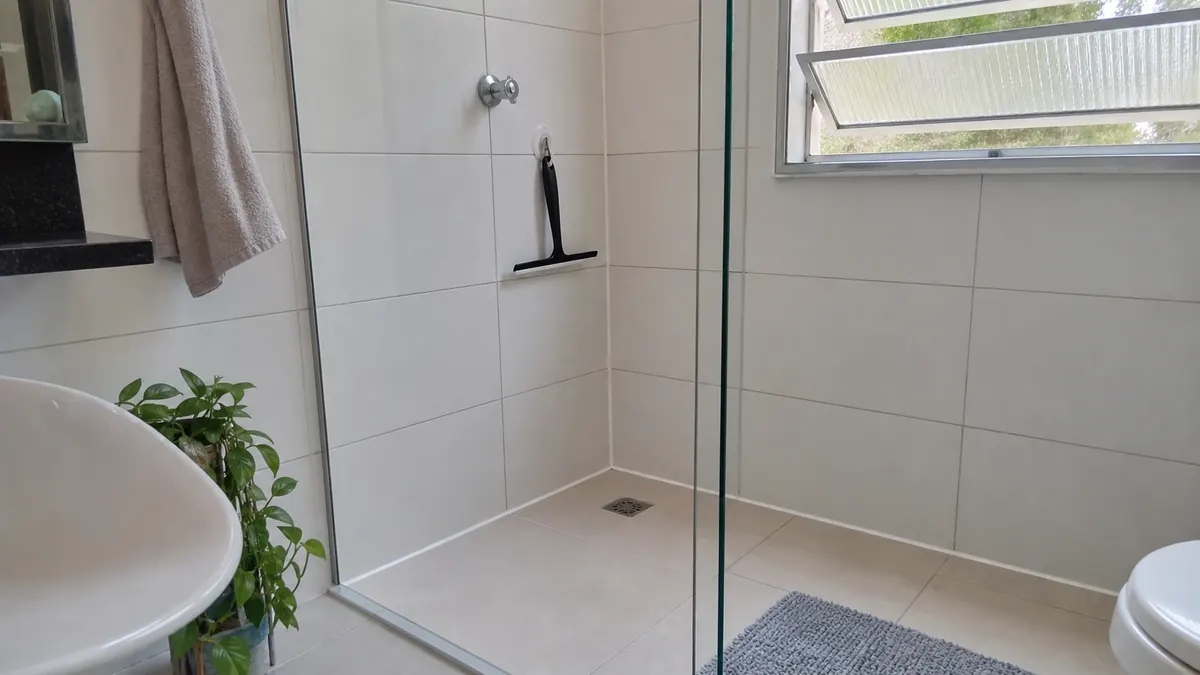 Box de banheiro pequeno com azulejo branco, rejunte limpo, silicone claro no canto, janela aberta e rodo pendurado.