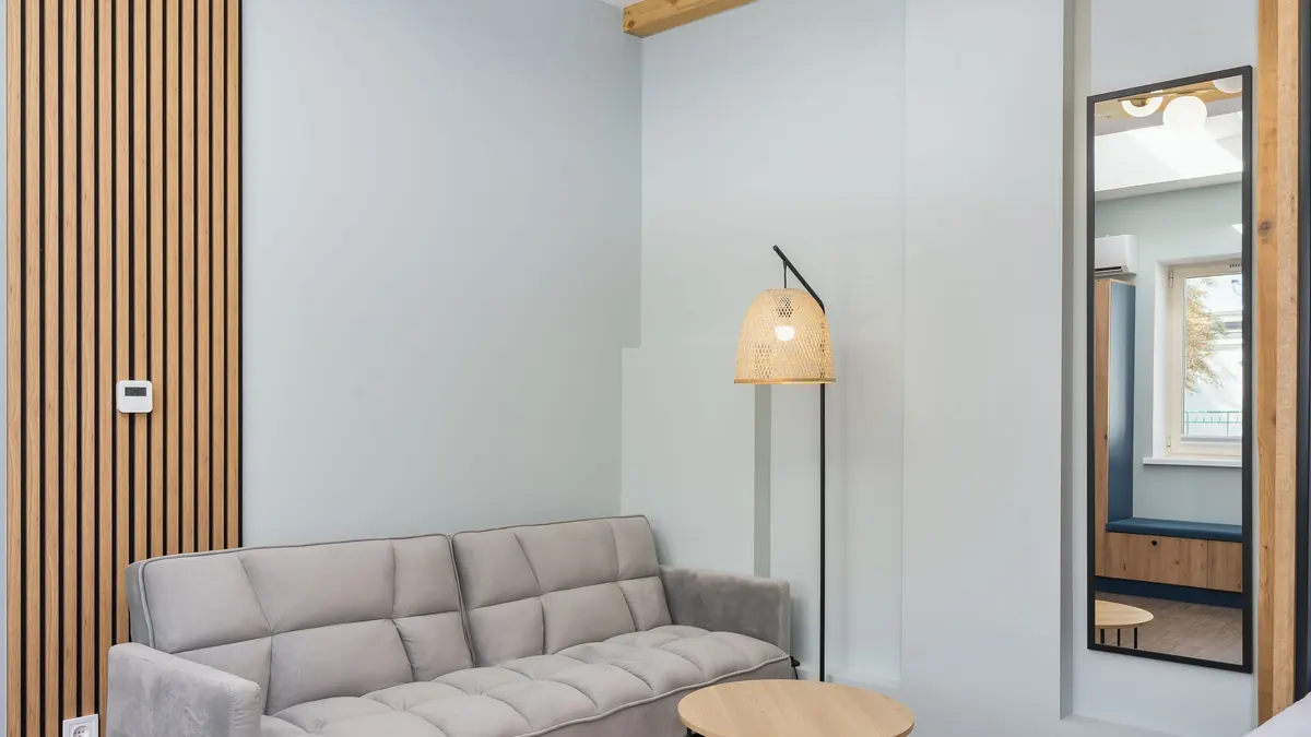 Sala com sofá cinza iluminada por abajur quente, cortina clara, madeira e parede em tom areia.