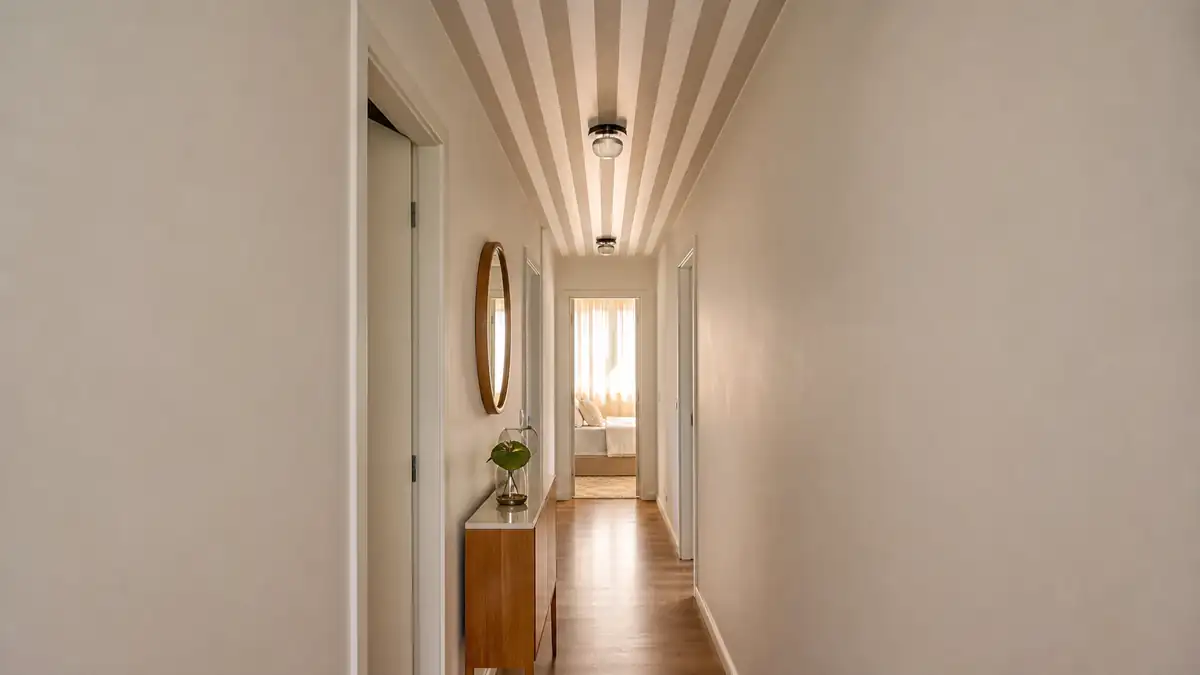 Corredor estreito de apartamento com teto listrado em tons claros guiando o olhar até o quarto iluminado.